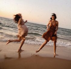 two women are running on the beach near the ocean at sunset or sunrise, one is wearing a brown dress