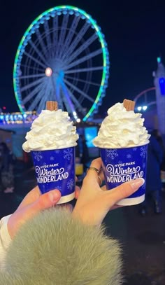 someone holding up two cups of ice cream in front of a ferris wheel at night