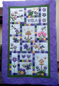 a purple and green quilted wall hanging on the side of a building with flowers