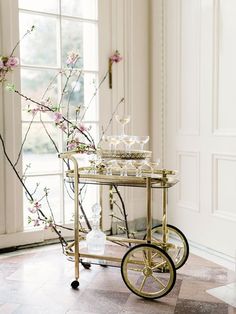 a gold bar cart with wine glasses on it in front of a window filled with pink flowers