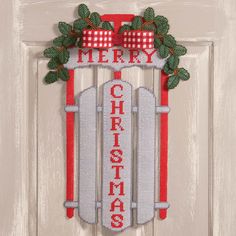 a cross stitch christmas door hanger hanging on a white door with holly wreath and bow