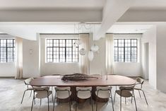 a large wooden table surrounded by chairs in an empty room with two windows on the wall