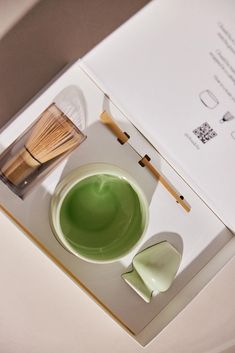 a green cup and whisk sitting on top of a white table next to an open book