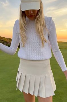 a woman in a white tennis outfit and hat is standing on the grass with her hands behind her back