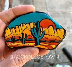 a hand holding a painted rock with a desert scene in the backgrounnd