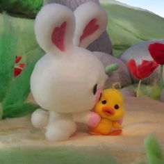 a small yellow chickling next to a white bunny in front of some rocks and flowers