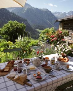 an outdoor table with food on it and mountains in the background is covered by flowers