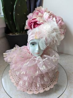 a white mannequin head with pink flowers in the background and a potted plant behind it