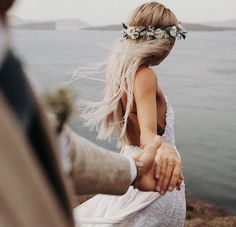 a man and woman holding hands near the water with flowers in their hair, while they hold each other's hand