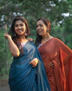 two women standing next to each other wearing sari and posing for the camera with trees in the background