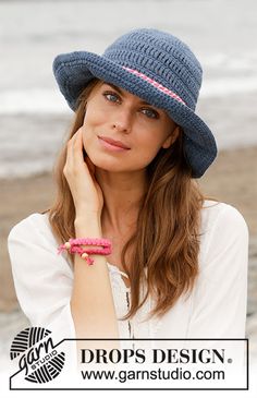 a beautiful young woman wearing a blue hat and pink bracelet sitting on the beach with her hand under her chin