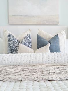 a white bed topped with lots of pillows next to a painting on the wall above it