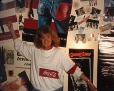 a woman standing in front of a wall covered with pictures and posters, holding her arms up