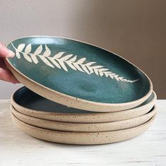 a hand is holding a green and white bowl on top of another dish that has leaves painted on it