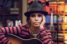 a woman wearing a hat and holding a guitar in front of a bookshelf