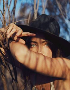 a woman wearing a hat and holding her hands up to her face while standing in the woods