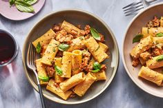 two plates of pasta with meat and basil on them next to a glass of red wine