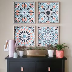 three paintings on the wall above a black cabinet with two planters and one potted plant