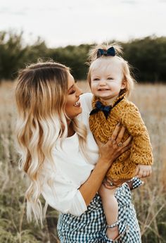 a woman holding a baby in her arms and smiling at the camera while standing in a field