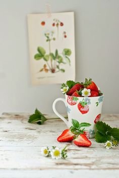 strawberries and daisies sit in a cup on a table next to a painting