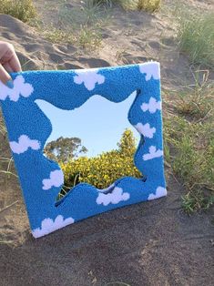 a hand holding up a blue and white frame with clouds on it in the sand