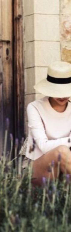 a woman wearing a white hat sitting on the ground next to purple flowers and looking at her cell phone