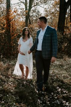 a pregnant woman in a white dress and a man in a suit are walking through the woods