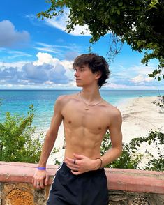 a shirtless young man standing next to a stone wall near the ocean with his hands on his hips