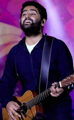 a man with a beard playing a guitar and singing into a microphone in front of a purple background