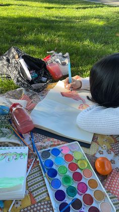 a woman laying on top of a lush green field next to a book and painting supplies