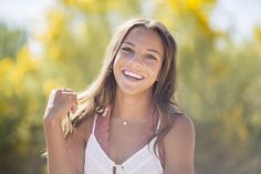 a beautiful young woman standing in front of trees smiling at the camera with her arms behind her back