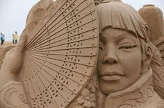 a sand sculpture of a woman holding an umbrella and looking at the camera with people in the background