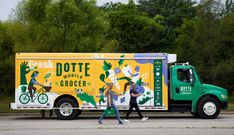 two people walking past a yellow and green truck on the side of the road with trees in the background