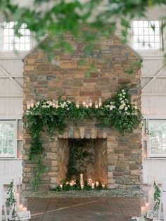 an indoor fireplace with candles and greenery on the mantle, surrounded by white chairs