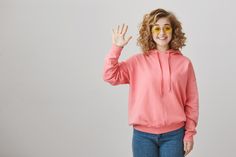 a woman with curly hair wearing a pink hoodie and yellow sunglasses is standing in front of a white background