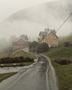 a rural road with houses on the side and fog in the air behind it, surrounded by mountains