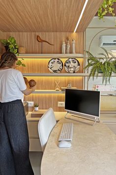 a woman standing in front of a desk with a computer on it next to a potted plant