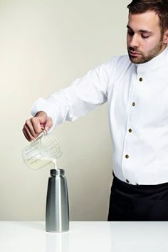 a man pouring something into a blender on top of a white table next to a silver shaker