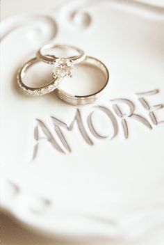 two wedding rings sitting on top of the word amore written in white paper with silver lettering