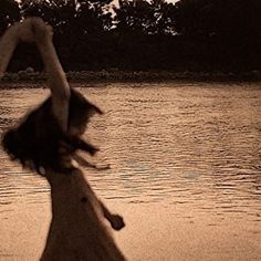 a woman standing in front of a body of water with her hands up to the sky