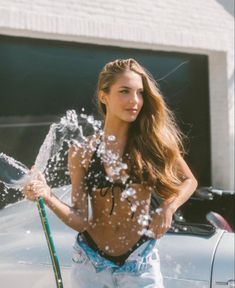 a beautiful young woman standing in front of a car holding a hose and spraying water on her face