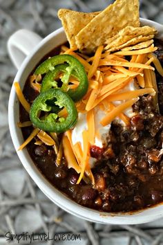 a white bowl filled with chili, cheese and tortilla chips next to a green pepper