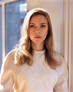 a woman with long blonde hair standing in front of a window wearing a white dress