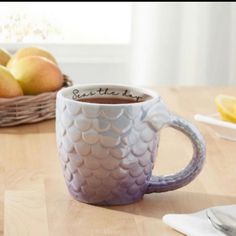 there is a coffee cup on the table with some fruit in the basket behind it