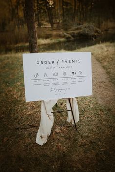 a white sign sitting on top of a grass covered field next to a forest filled with trees