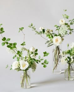 three clear vases with white flowers and greenery in them on a gray background