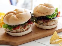 two chicken burgers with lettuce and tomato on a cutting board next to chips