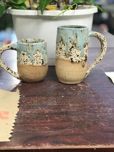 two coffee mugs sitting next to each other on top of a wooden table in front of a potted plant