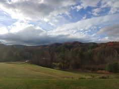 an open field with trees in the background and clouds in the sky over mountains behind it