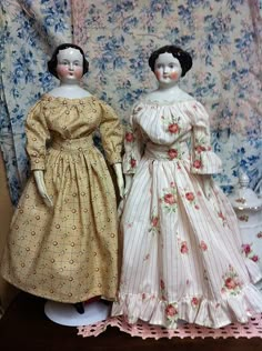 two dolls are standing next to each other on a wooden table in front of floral wallpaper